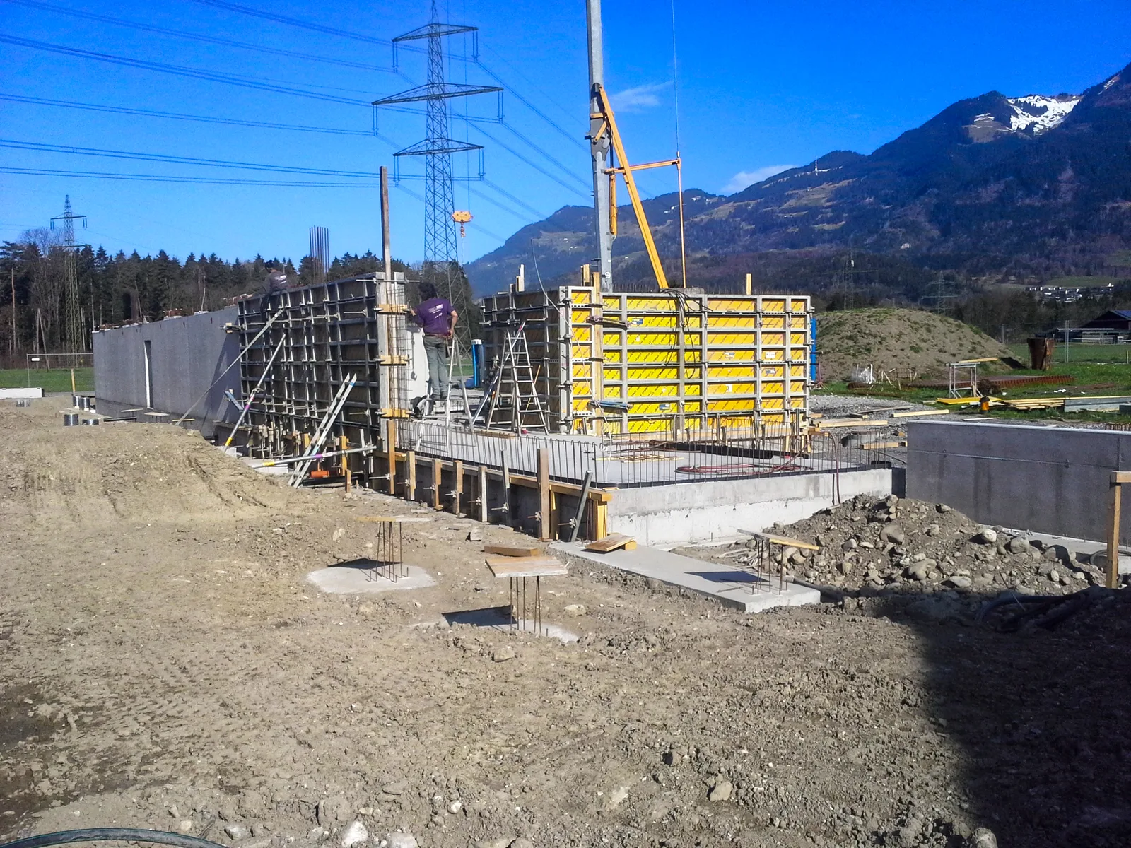 Baustelle mit hoher gelber Stützenschalung und Gerüst, dahinter ein Kran, Strommasten und Berge