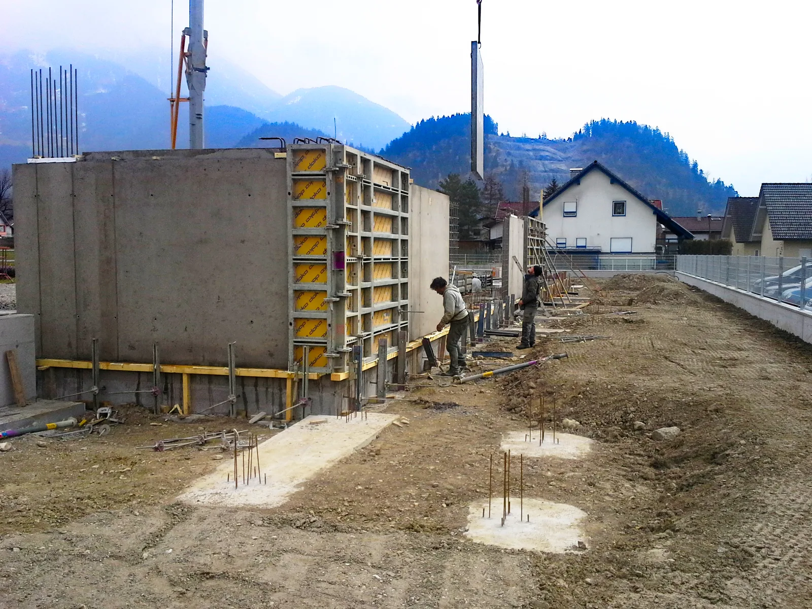 Aufgestellte Schalungswand auf einem Fundament, zwei Arbeiter daneben, im Hintergrund ein Dorf am Hang
