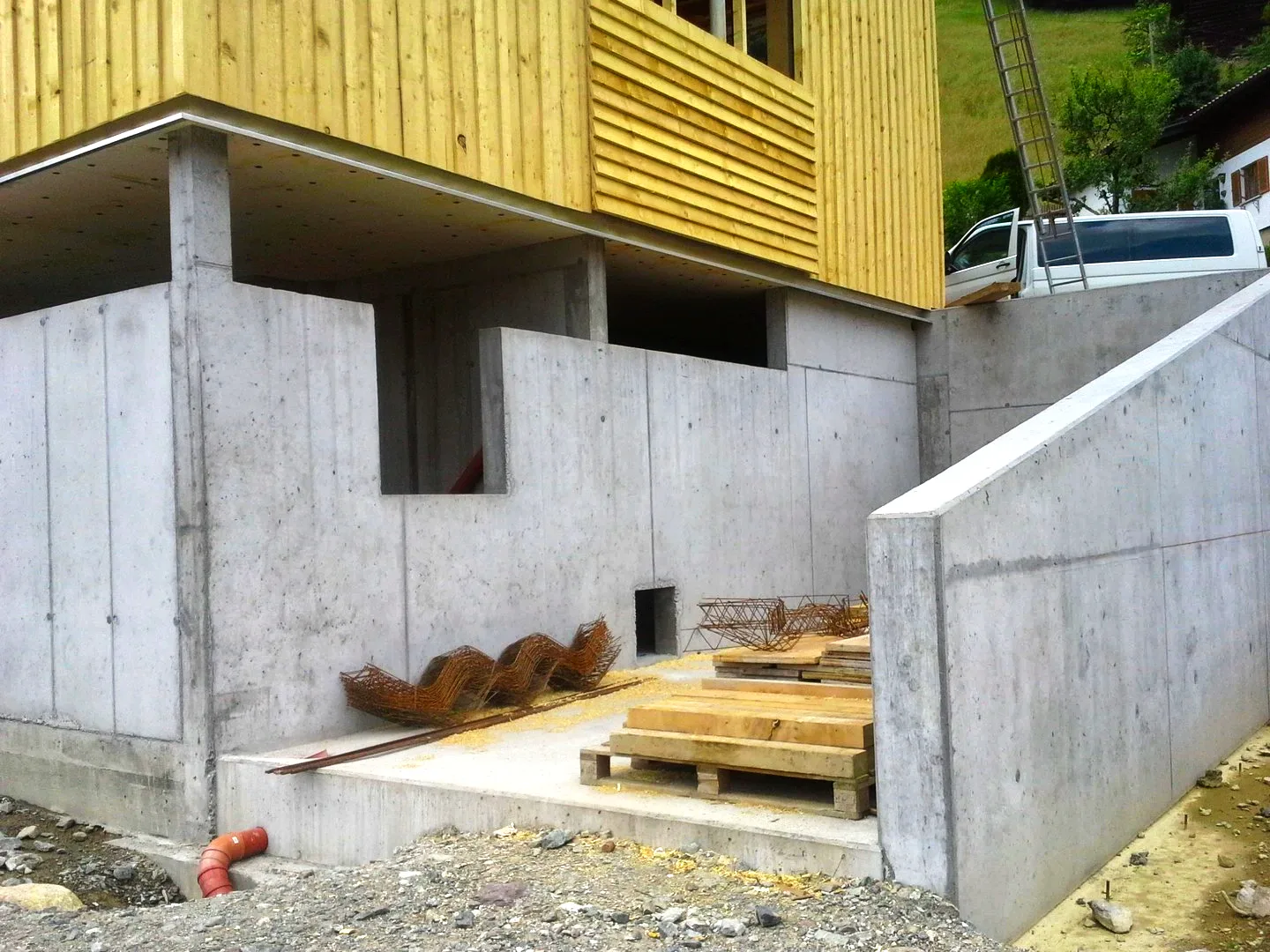 Sichtbeton-Sockel mit Stützmauer unter einer hellen Holzfassade, davor frisch verlegte Bretter