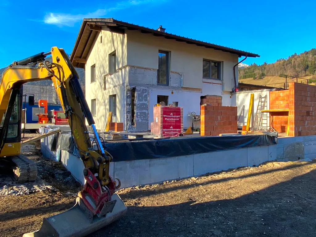 Minibagger vor einem älteren Wohnhaus, daneben ein frisch aufgemauerter Anbau mit Abdichtungsbahn