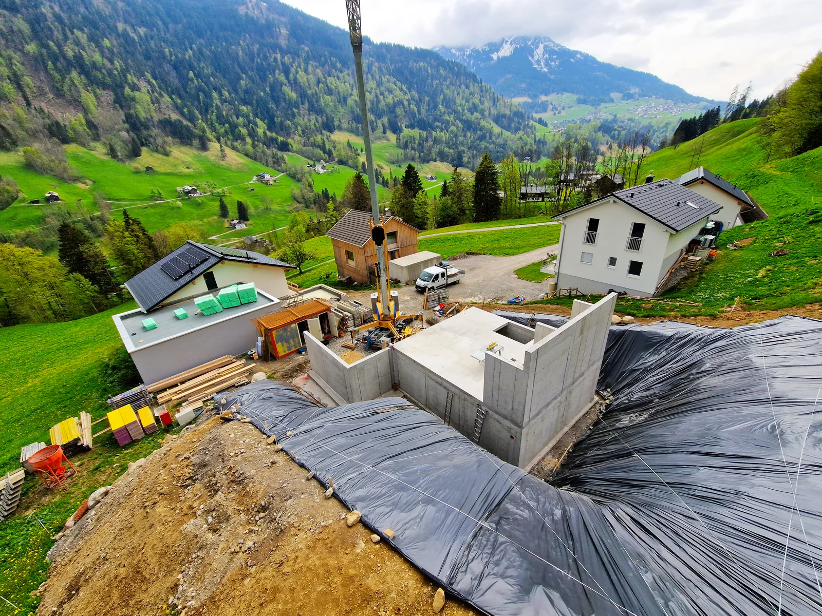 Blick von oben auf einen Rohbau mit schwarzer Abdichtungsfolie, ringsum Wiesen und bewaldete Hänge