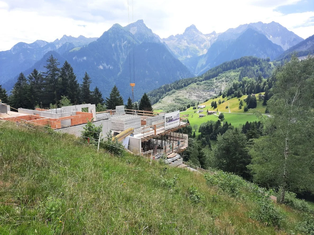 Rohbau am Hang mit aufgestelltem Kran, dahinter ein weites Tal und schneebedeckte Gipfel