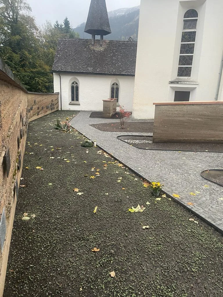 Gepflasterter Vorplatz entlang einer weißen Kirchenwand, daneben eine niedrige Natursteinmauer