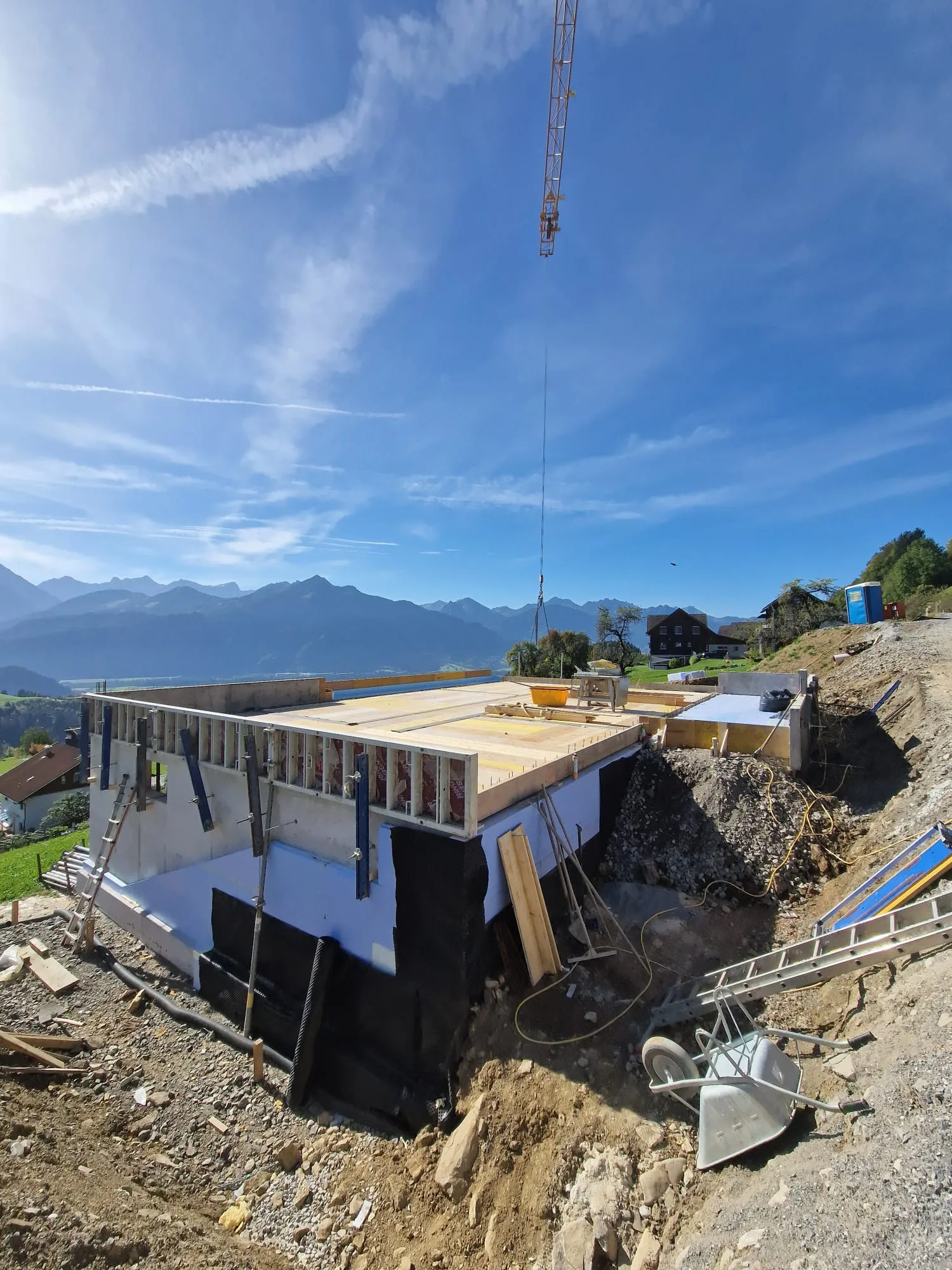 Kellerrohbau mit aufgestellter Schalung und Baukran, dahinter ein breites Alpental